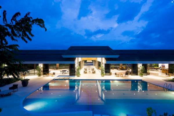 An illuminated outdoor swimming pool with poolside seating leads to a modern building under a twilight sky.
