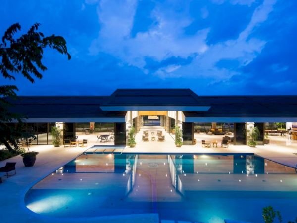 An illuminated outdoor swimming pool with poolside seating leads to a modern building under a twilight sky.