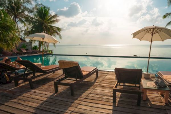 A serene poolside scene overlooking a calm ocean. Deck chairs and umbrellas are set on a wooden deck, surrounded by palm trees under a bright sky.