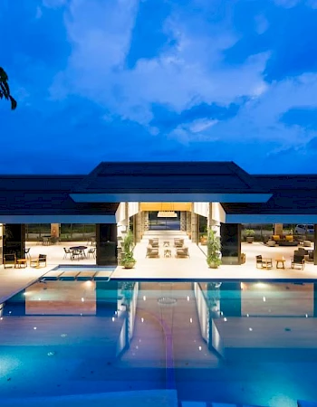 The image features a large, illuminated swimming pool in front of a modern, open-concept building under a twilight sky, surrounded by greenery.