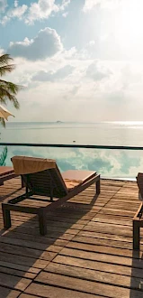 Outdoor pool scene with lounge chairs, umbrellas, palm trees, and a view of the ocean under a partly cloudy sky.