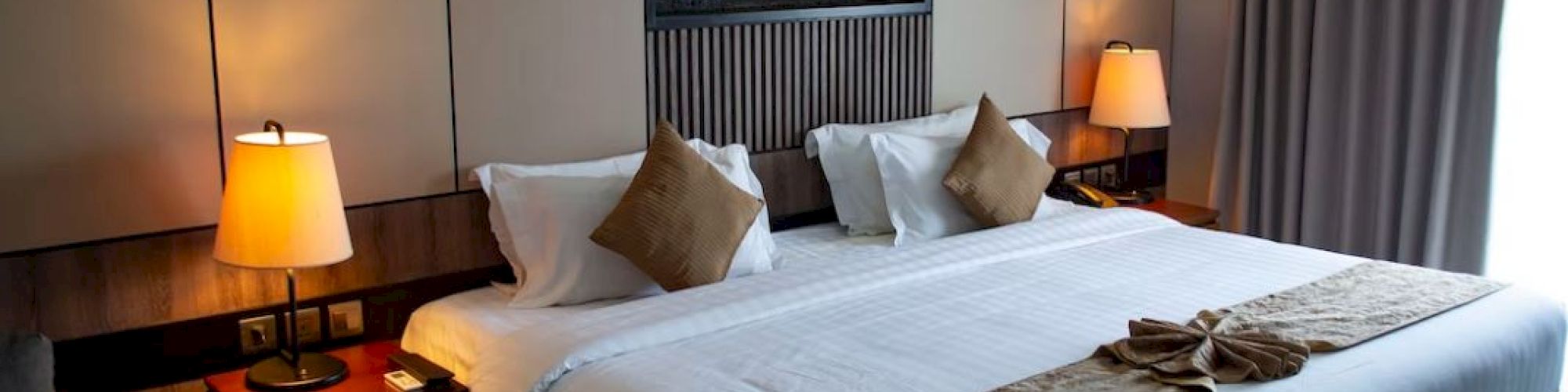 A modern hotel room features a large double bed, two bedside lamps, and a wall-mounted fireplace. The room is well-lit and elegantly decorated.
