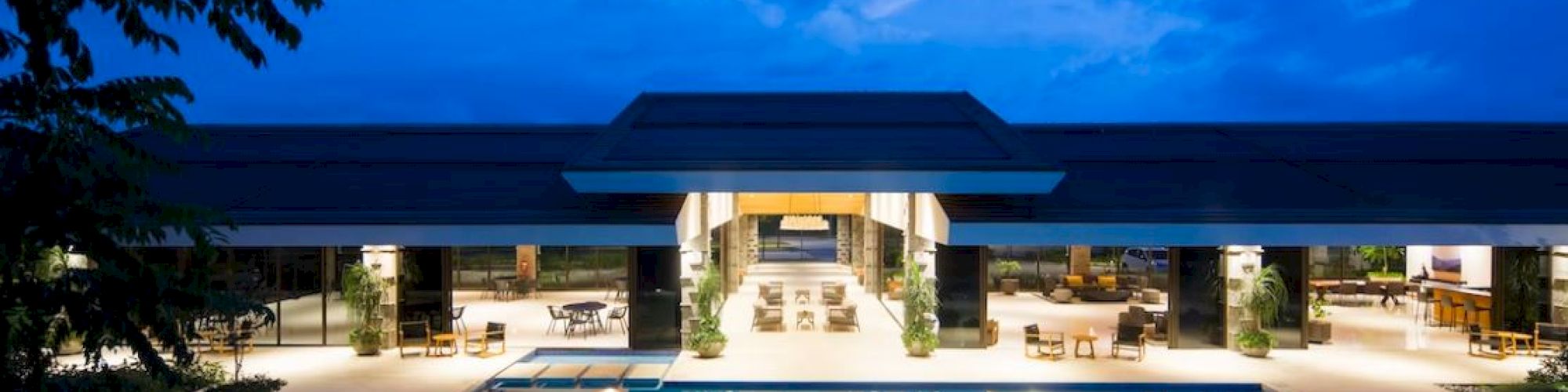The image shows a modern building with an illuminated swimming pool and spacious open seating area at dusk, under a vibrant blue sky.