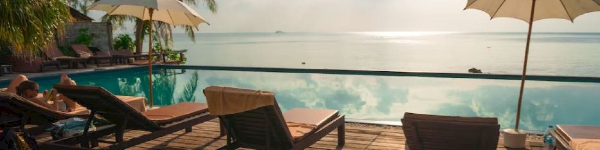 Sun loungers and umbrellas by a calm pool overlooking the sea, with palm trees and a bright, cloudy sky in the background.
