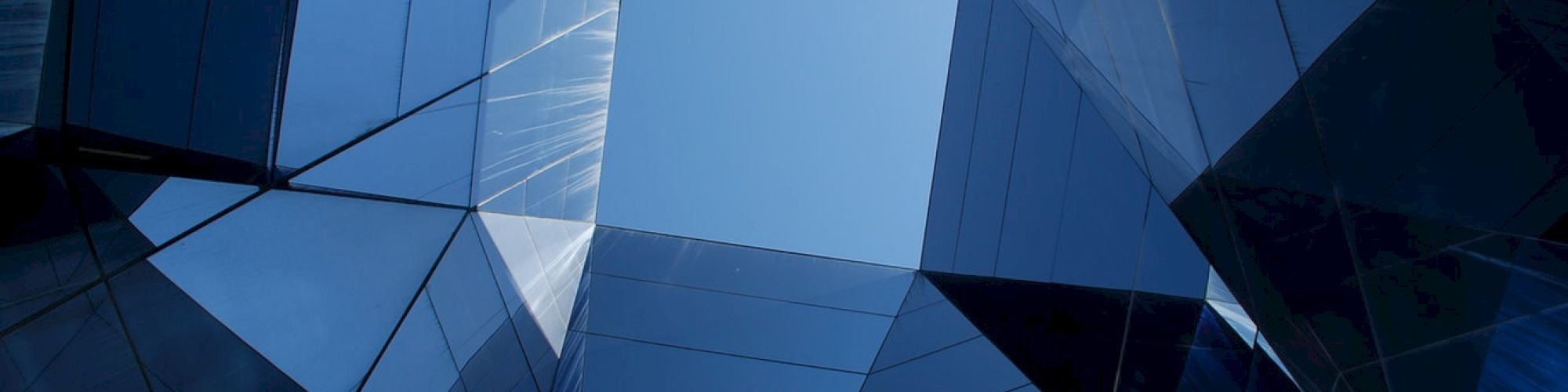 The image shows an upward view of a modern building's glass structure, creating geometric patterns and reflecting the blue sky in the center.