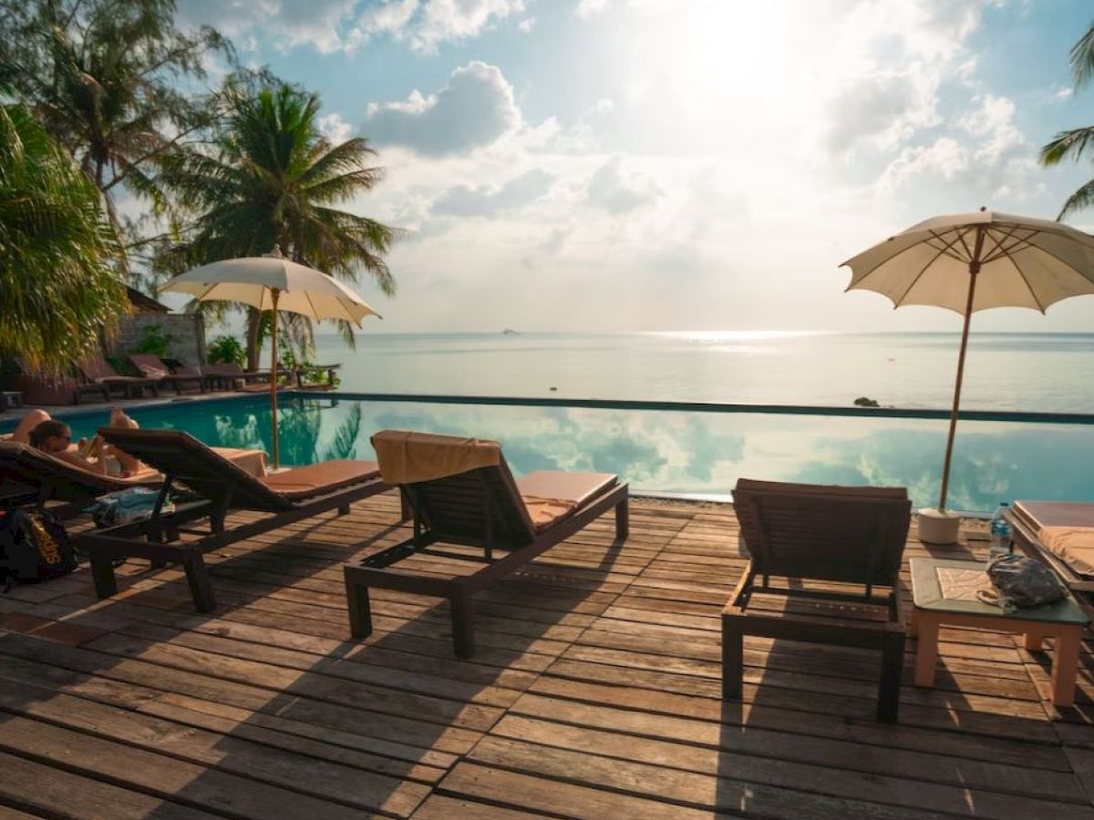 Poolside deck with lounge chairs, umbrellas, and a scenic ocean view, surrounded by palm trees and bathed in sunlight.