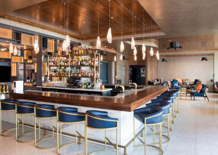 A modern bar with a row of chairs, a well-stocked shelf of liquor bottles, hanging lights, and a mix of tables and seating in the background.