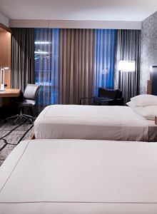 A modern hotel room with two beds, a wall-mounted TV, desk, and floor-to-ceiling windows with curtains. The room is well-lit and neatly arranged.