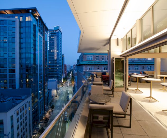 A modern cityscape view from a high-rise balcony with tables and chairs, overlooking the street and buildings at dusk, highlighting urban living.