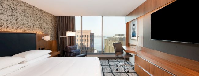 A modern hotel room features a king bed, a large flat-screen TV, a desk, and floor-to-ceiling windows offering a cityscape view.