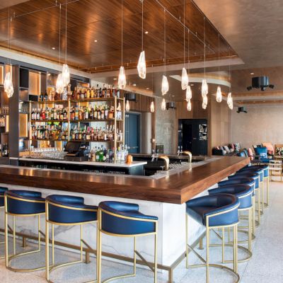 The image shows a modern bar with several blue bar stools, a well-stocked bar, hanging lights, and a seating area in the background.