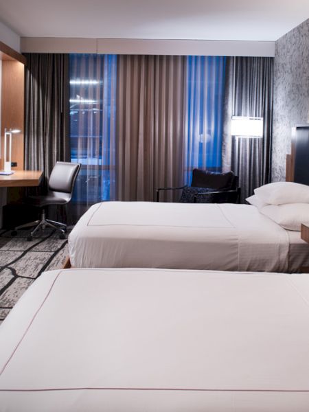 A modern hotel room with two beds, a TV, a desk area, and contemporary lighting and decor. The room appears clean and comfortable.