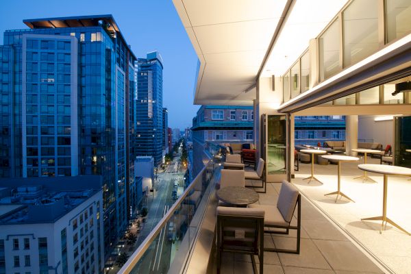 A modern balcony with seating overlooks a city street lined with tall buildings at dusk, and through the open glass wall, you can see a modern room.