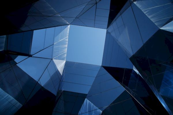 This image shows a geometric view of glass buildings from below, with a blue sky visible through the angular structures formed by their reflections.