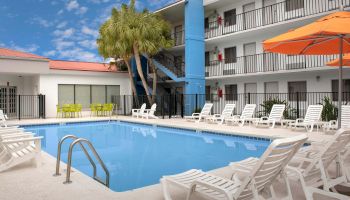 The image shows an outdoor pool at a hotel or resort, with white lounge chairs and an orange umbrella, surrounded by a multi-story building.