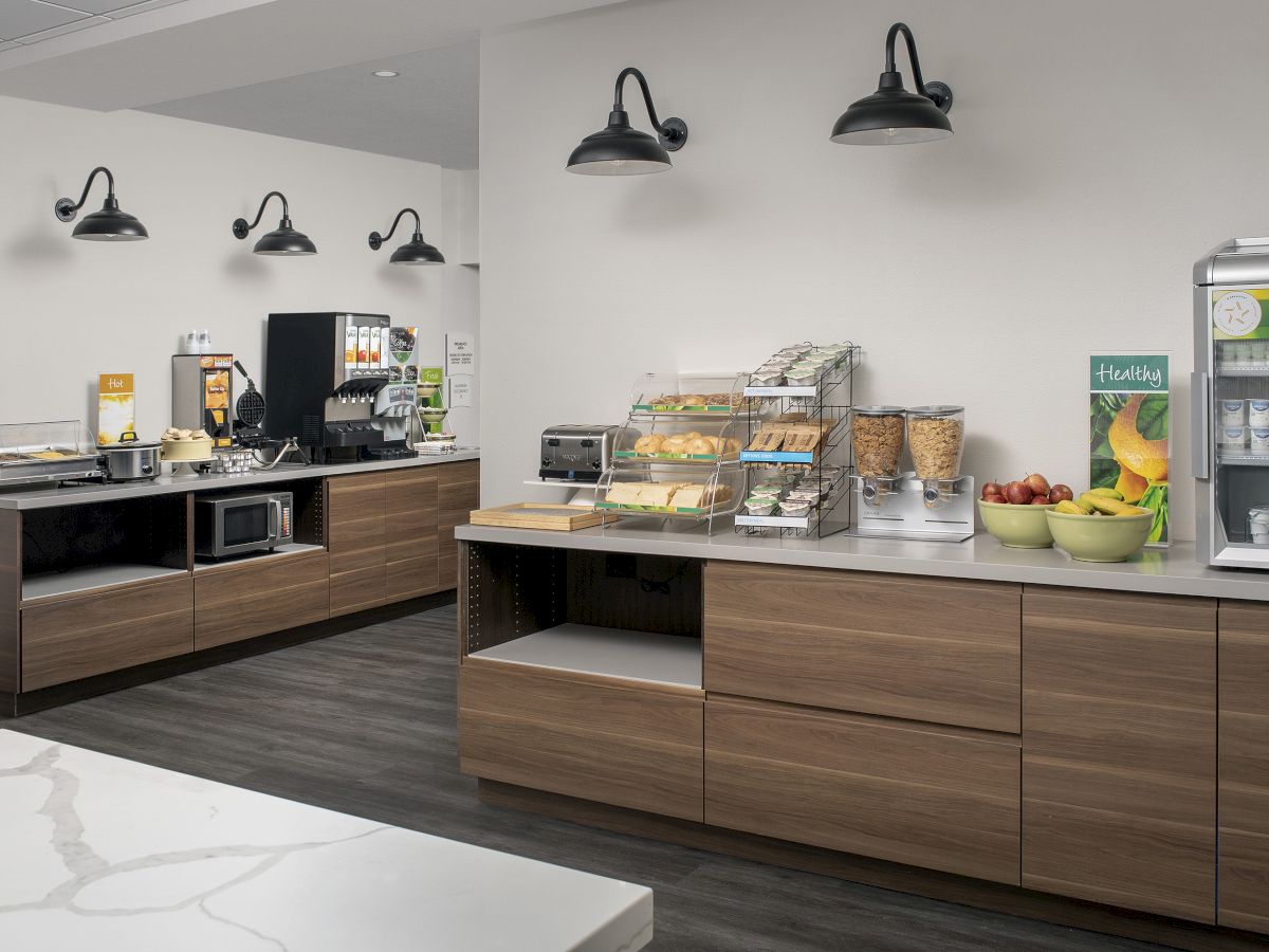 The image shows a modern snack and breakfast bar with coffee machines, a microwave, cereals, fruits, and refrigerated drinks.