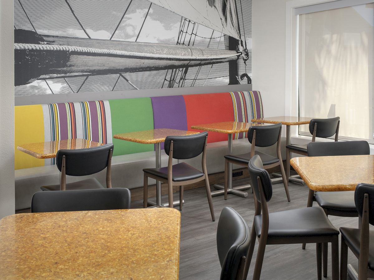 The image shows a modern cafeteria with colorful, striped booth seating, wooden tables, black chairs, and a black-and-white mural on the wall.