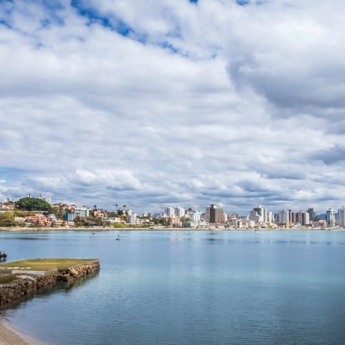 The image shows a coastal cityscape with buildings along the shoreline, a partly cloudy sky, and calm water with a small pier on the left side.