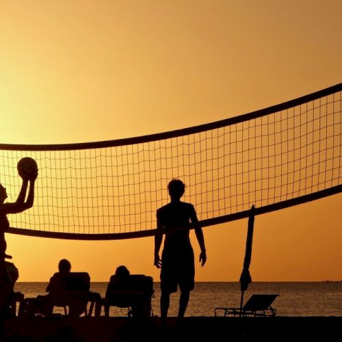 People are playing volleyball on a beach at sunset, while other individuals are sitting and relaxing by the shore.