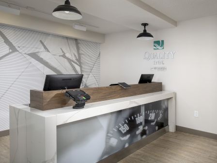 A reception desk with two computers is in a modern, well-lit area. The wall has a design with a 