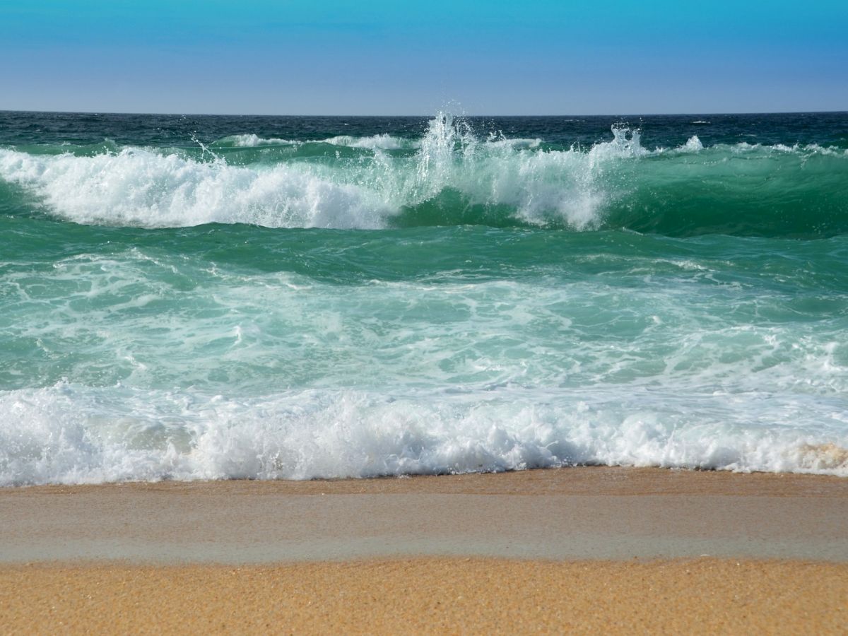 The image shows a beach with waves crashing onto the shore, clear blue skies, and a beautiful ocean view.