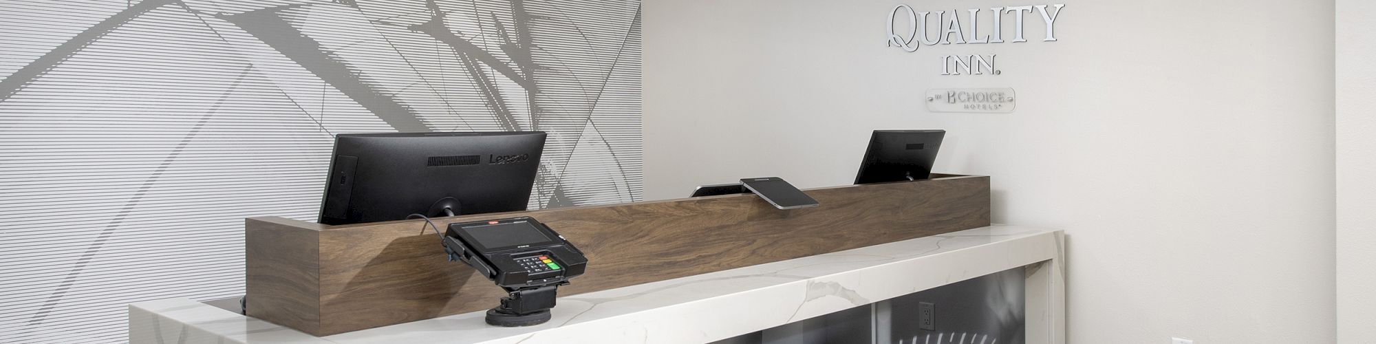 This image shows a modern hotel reception desk with two computers, card readers, and an abstract wall design with the hotel logo displayed on the wall.