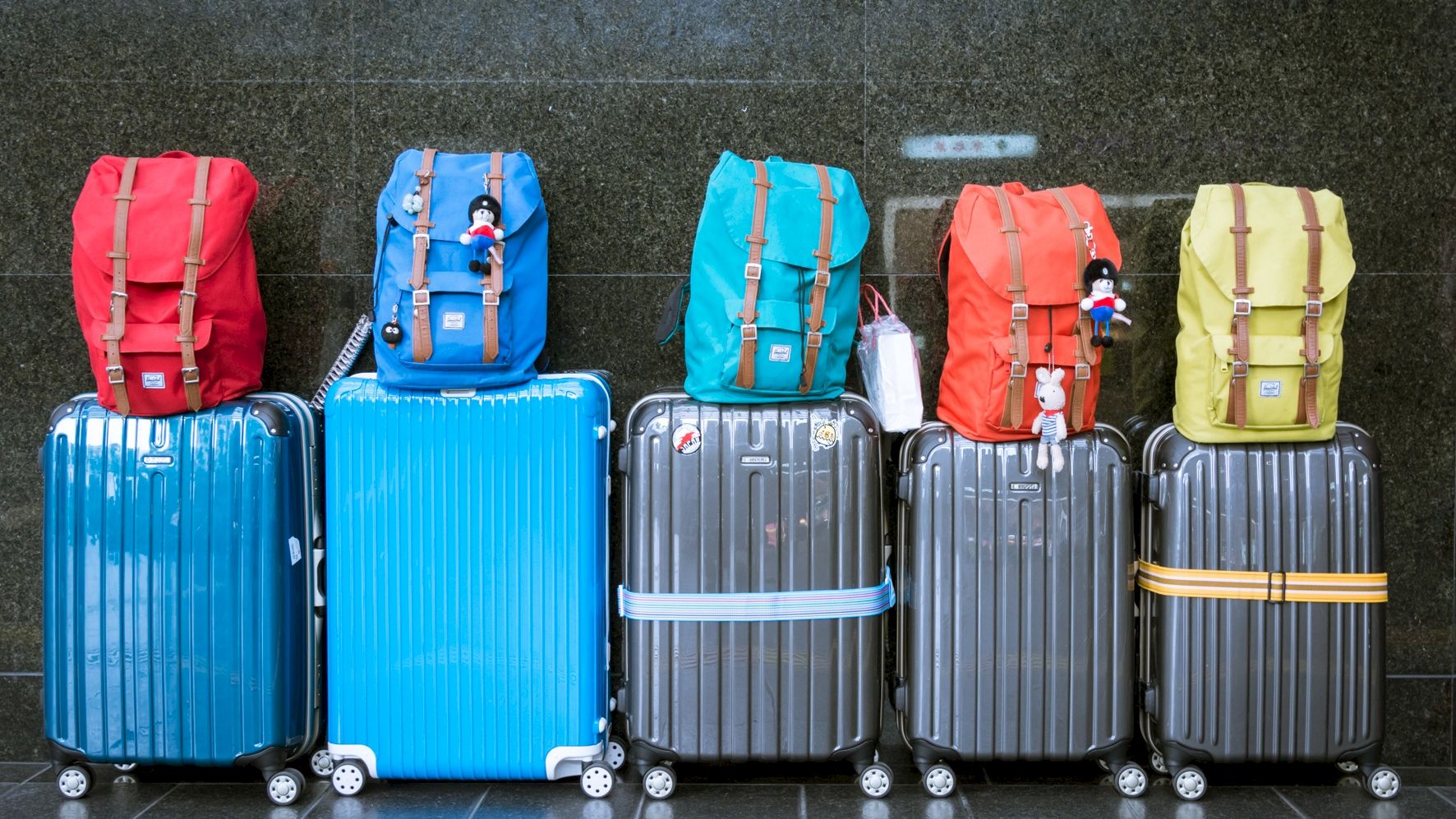 The image shows five suitcases, each with a backpack on top, arranged in a row against a dark wall. The suitcases are blue and gray.