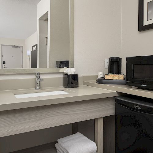 The image depicts a bathroom area with a sink, mirror, towel, coffee maker, microwave, and small fridge underneath a granite countertop.