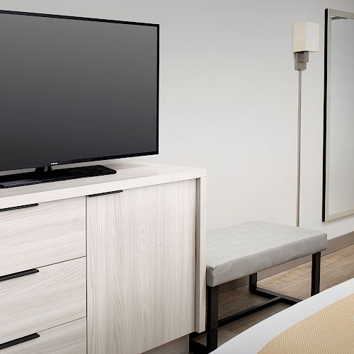 The image shows a minimalist hotel room with a flat-screen TV on a light wood dresser, a wall-mounted mirror, a bench, and a bed corner.