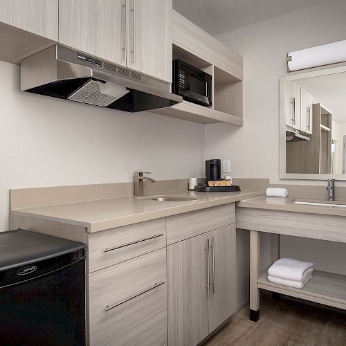 A small kitchenette with light wood cabinets, a mini-fridge, sink, microwave, coffee maker, and a countertop with a mirror on the right side.