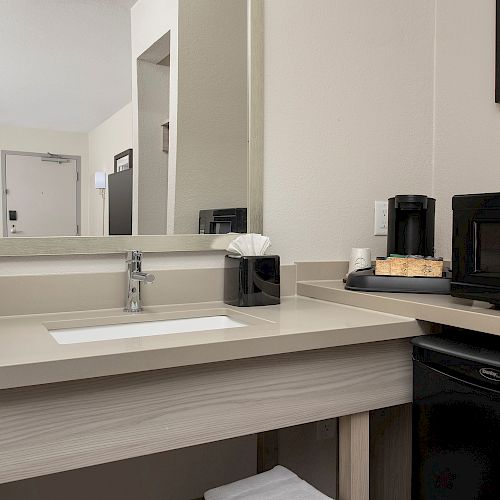 The image shows a hotel room kitchenette area with a sink, mirror, microwave, coffee maker, and a small refrigerator on a countertop.