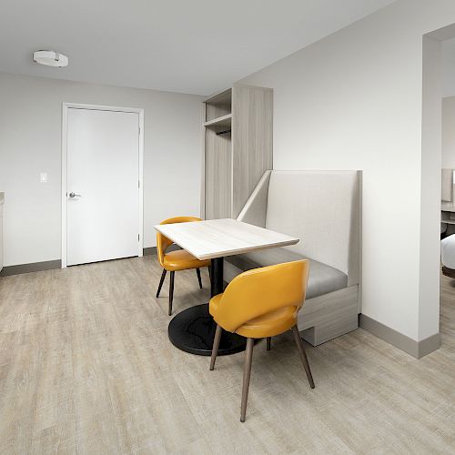 This image shows a modern hotel room with a small dining area, kitchenette, two yellow chairs, and another room with a bed visible through an open door.