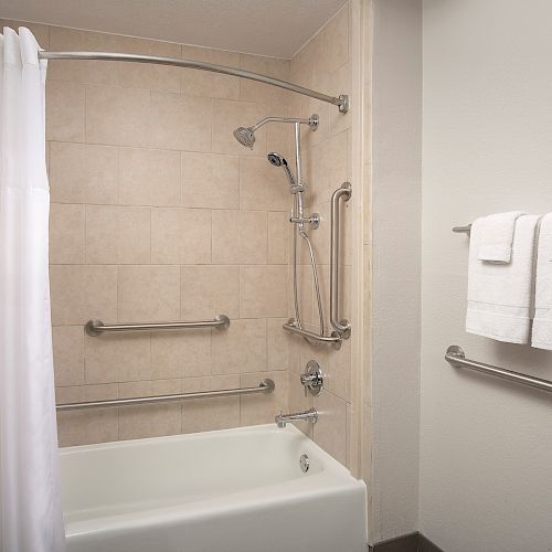 This image shows a bathroom with a bathtub, a showerhead, grab bars, a white shower curtain, and a wall with white towels hanging on towel racks.