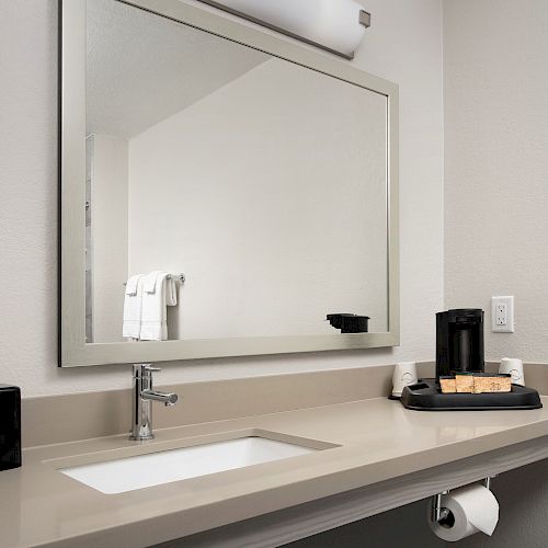 A bathroom sink with a large mirror, a tissue box, a toilet paper roll, and a coffee maker with amenities on the countertop.