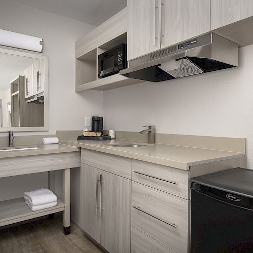 The image shows a modern kitchenette with light wood cabinets, a sink, a microwave, a coffee maker, a mini fridge, and a mirror above the sink.