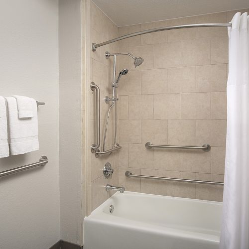 A bathroom with a tiled shower and tub, white shower curtain, multiple grab bars, and white towels hanging on a rack by the wall.