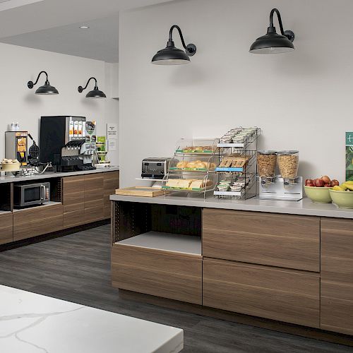 A well-organized breakfast area with various food items, a fridge, microwave, and cereal dispensers on wooden counters with modern lighting.