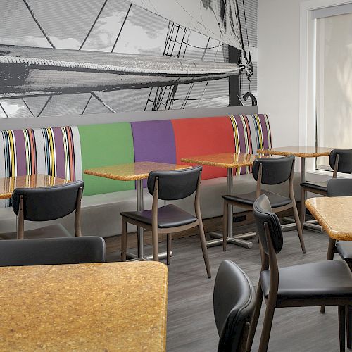 The image shows a modern dining area with colorful booth seating, black chairs, and wooden tables, featuring a large mural of a sailboat.