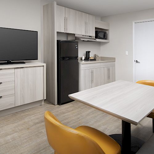 The image shows a small kitchen area with a refrigerator, microwave, countertop, TV on a stand, and a table with two yellow chairs, ending the sentence.