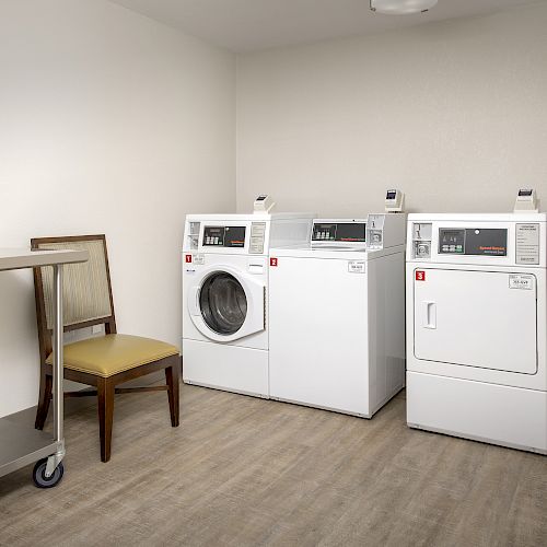 The image shows a laundry room with a washing machine, two dryers, a chair, and a metal cart.