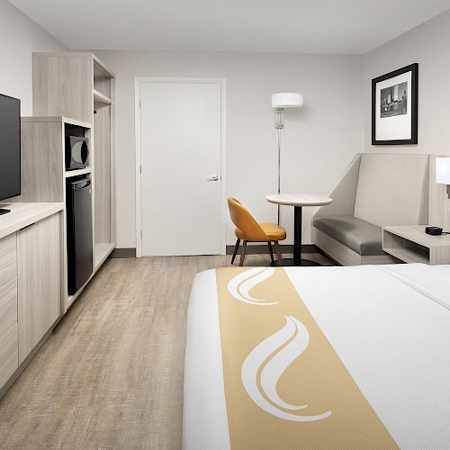 A hotel room with a bed, TV, mini-fridge, microwave, table, chairs, and modern decor, featuring light-colored furnishings and flooring.