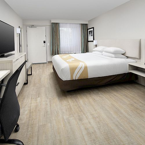 A modern hotel room with a bed, desk, chair, TV, and a small table with two chairs, featuring minimalist decor and wooden flooring.