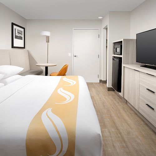 A modern hotel room features a bed, TV, microwave, mini-fridge, desk, and chair with minimalistic decor and neutral tones.