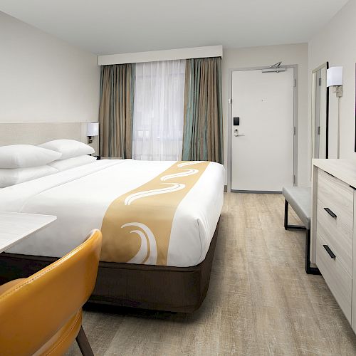 The image shows a modern hotel room with a neatly made bed, TV, and light-colored furniture. There's also a chair and a small table.