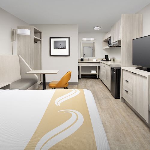 This image shows a modern hotel room with a bed, desk, chair, TV, kitchenette, and neutral decor.