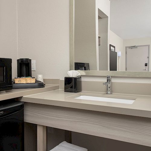 A bathroom countertop with a sink, mirror, black microwave, mini-fridge, coffee maker with supplies, tissue box, and a rolled towel.