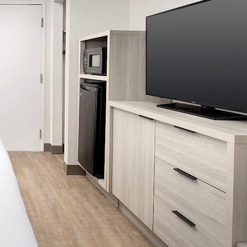 The image shows a hotel room with a bed, a TV on a light-colored cabinet, a microwave, and a small fridge by a closed door.