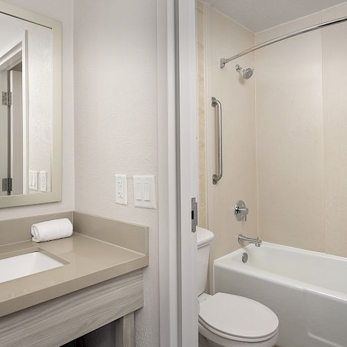 The image shows a modern bathroom with a sink and mirror on the left, and a bathtub with a shower curtain and toilet on the right, with neutral tones.