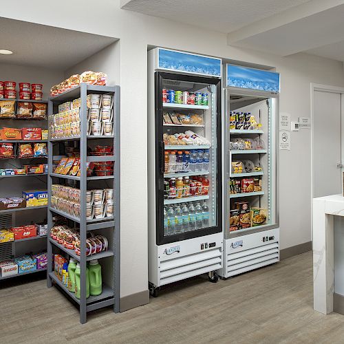 The image shows a small convenience store with shelves stocked with snacks and refrigerated beverages. A counter with a register is on the right side.