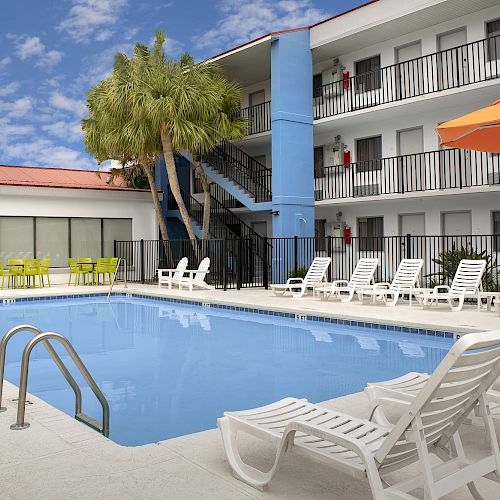 An outdoor swimming pool area with lounge chairs, an orange umbrella, and a three-story building in the background with balconies.